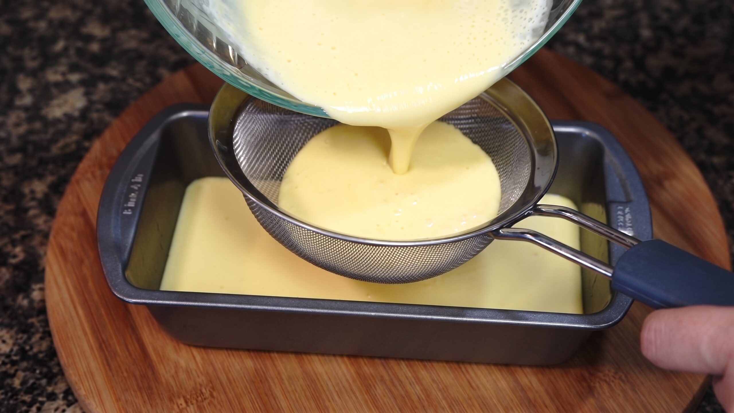 Mango custard mixture being poured through a fine-mesh strainer into the caramel-lined container